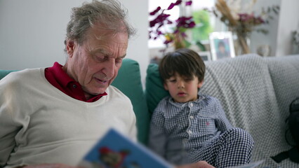 Grandfather reading a colorful storybook to his young grandson, both sitting on a cozy couch, deeply engaged in the tale, creating a moment of bonding and shared learning at home