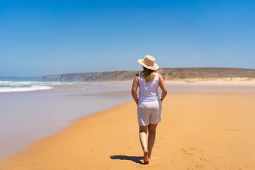 Woman wearing light clothes for hot weather and sun hat walking towards cliffs on sandy beach on summer day. Bordeira praia, Portugal. Relax and calm on beach. Back view