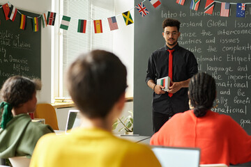 Young male teacher standing by blackboard in school classroom and talking to students, copy space