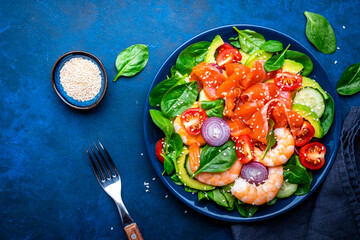 Healthy food salmon salad for keto diet with shrimp, avocado, spinach, cucumber, tomato, cashew, sesame seeds. Blue background, top view
