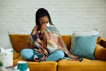 Young black woman with flu virus sneezing in tissue at home.