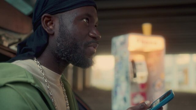 African American man wearing durag scrolling through social media on his mobile phone, smiling and telling something while standing by car with payphone booth in background under bridge