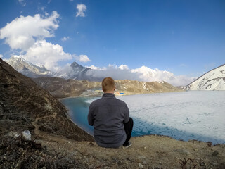 Naklejka premium Rear view of male person sitting by ice partly covered Gokyo lake and Gokyo village in Gokyo mountain valley in Nepal in Himalayas on height 4750 meters in a sunny day. Travel and hiking theme.