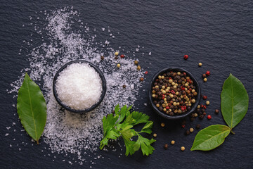 Sea salt and peppercorns in small  dishes. Bay laurel leaves and parsley leaves on black...
