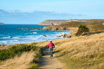 nice senior woman cycling with her electric mountain bike at the rocky emerald coast  of Brittany, France next to Cap Frehel