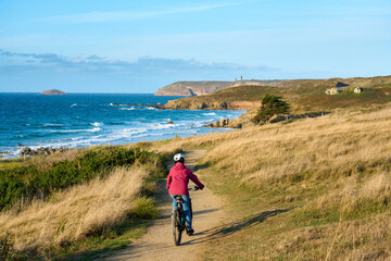 nice senior woman cycling with her electric mountain bike at the rocky emerald coast  of Brittany, France next to Cap Frehel