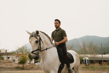 Portrait of a young rider on his white horse in an equestrian centre with mountains in the...