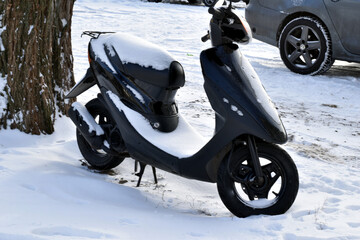 A scooter covered in white snow stands near a tree.