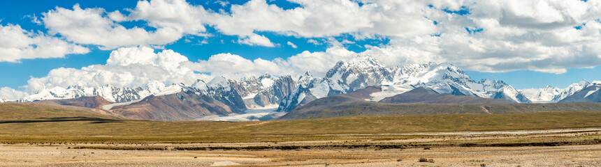 Yellow fields stretch out in front of rocky mountains,tops of which are covered with snow.The...