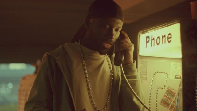 Black man in durag having emotional conversation on retro payphone covered in stickers and illuminated by warm light in evening, people walking on street under overpass behind him