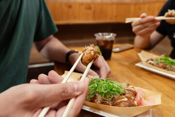A man is eating a takoyaki ball with his chopsticks in a bar with friends. A popular street food in Osaka, Japan