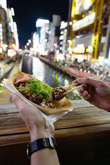 Obraz premium A tourist is eating a takoyaki ball with chopsticks, popular street food at Dotonbori canal in Osaka, Japan. It's a summer night, the lights of billboards are reflexing at the water in the background