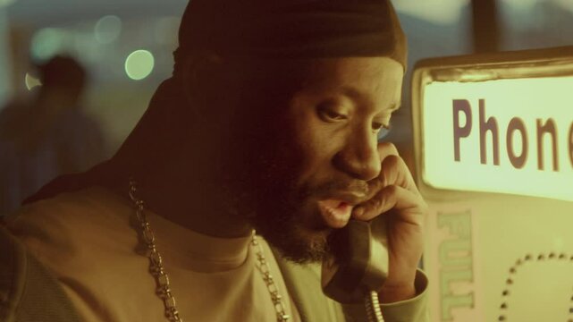African American man wearing silver necklace and durag having intense conversation on street payphone illuminated by warm light under city bridge in the evening. Close-up view