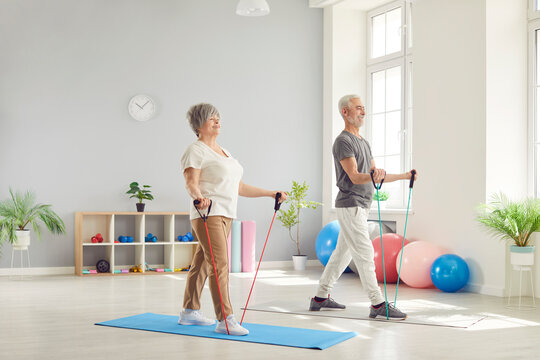 Smiling mature senior man and woman doing stretching exercises with rubber band standing in gym. Happy elderly people doing workout in rehabilitation center for health. Sport and fitness concept.