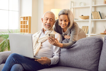 Senior family couple is enjoying leisure time together at home. They are sitting on the sofa with a laptop and mobile phone, engaging in online entertainment and leisure activities.