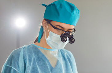 Headshot serious skilled woman surgeon doctor, wearing face mask, safety glasses, loupes, blue...