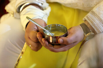 A person holding a small metal cup with a spoon, likely part of a traditional ceremony or offering, symbolizing purity and blessings in a sacred ritual.