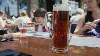 Large glass of dark amber beer with a frothy head on a wooden table at an outdoor cafe, showcasing the enjoyment of a refreshing drink in a social and relaxed setting