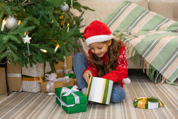 Christmas spirit. Happy little smiling girl opening gift box sitting on the floor near the Christmas tree. Happy girl unwrapping christmas present looking in box. Holidays and celebrations concept