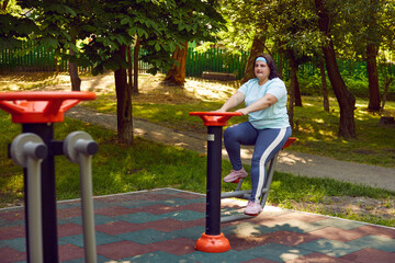 Overweight fat woman doing sport exercises on bike simulator outside in summer park. Plump young...