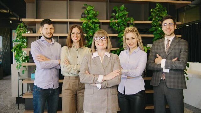 A group of businessmen headed by an elderly experienced female director confidently stands in a modern office, demonstrating ambition and cohesion in teamwork.