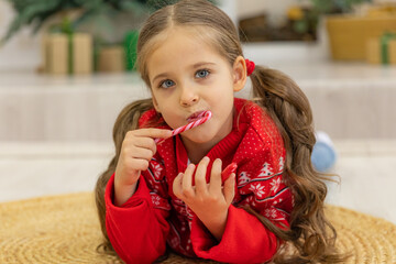 Happy little girl with a lollipop near Christmas tree at home. Little girl with sweet candies on Xmas eve. Family, tradition, celebration concept. Candy canes in shape of heart in hand child