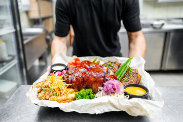 close-up of a plated meal held by a person in a kitchen, featuring roasted meat with garnishes and sauces. The dish is presented on a white plate, highlighting the vibrant colors and textures of food