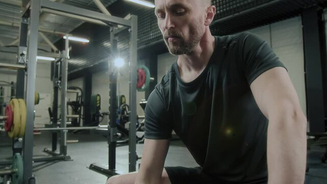 Tilt up shot of man in sportswear sitting on gym bench doing one arm dumbbell curl exercise during intense weight training