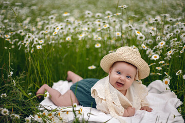 Fototapeta premium beautiful chubby little girl infant baby lying in a field with daisies in a hat on a summer afternoon in nature