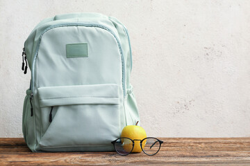 Green school backpack with apple and eyeglasses on wooden table near light grunge wall