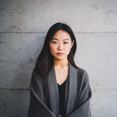 Fototapeta premium A portrait of a Japanese woman with a grey sweater standing against a concrete wall 