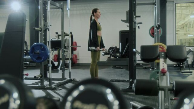 Full length shot of young fit woman in sportswear standing in well-equipped gym and performing barbell upright rows during weight training session