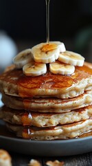 Stack of pancakes topped with banana slices and drizzled with syrup, served on a plate with golden brown texture and sweet toppings.