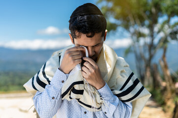 Young Jewish Man Outdoors with Tallit and Kipa, Passing Tzitzit Over His Eyes During Shacharit...