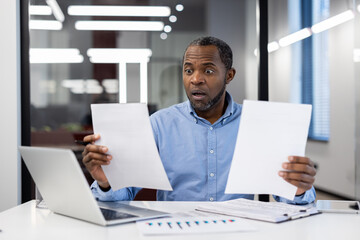 Businessman seated at office desk holding paperwork with surprised expression. Laptop open in front. Scene conveys shock, unexpected news, or financial report review in professional setting.