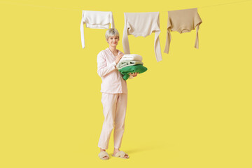 Beautiful young woman with stack of clean clothes and laundry hanging on rope against yellow background