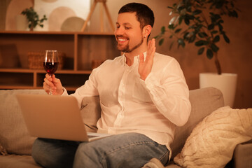 Obraz premium Handsome man with wine and laptop having online date on sofa at home in evening