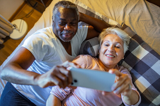 Happy diverse senior couple taking selfie in bed at home - Powered by Adobe