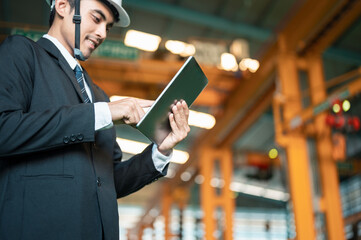 engineer, technician, architect, checking, distribution, manufacturing, hard hat, construction, safety, machinery. A man in a suit is using a tablet while standing in a large industrial building.