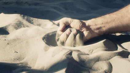 Close-up of a hand grasping and squeezing fine sand, creating ripples and shadows on a sun-drenched beach. The image captures the tactile sensation of sand slipping through fingers.
