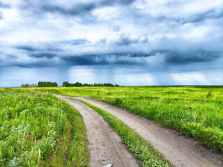 A dirt road winds through a grassy field, with a storm brewing in the sky overhead
