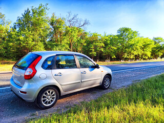 A silver compact car is parked beside a tree-lined road under clear skies