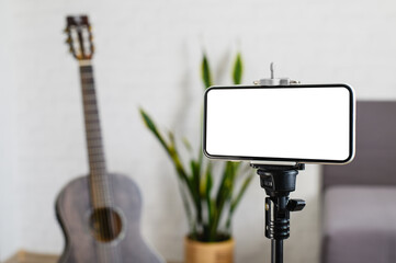 Online guitar lesson. Smartphone on a stand with a white display against the background of a guitar in a room. Selective focus on the smartphone.