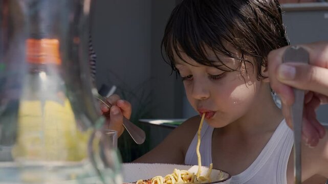 Young boy slurping a long noodle while eating spaghetti, using a fork to twirl the pasta and enjoying his meal outdoors at a table during the day