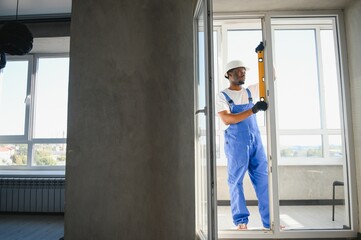 Workman in overalls installing or adjusting plastic windows in the living room at home