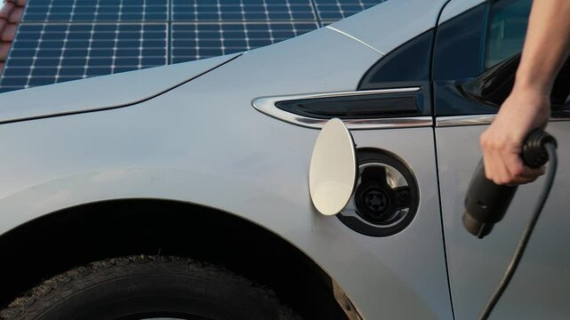 Zoom out shot of male hand disconnects power cable supply from EV or electric car on the solar panels background. Unrecognizable man unplugs an electric auto or PHEV from photovoltaic charging station