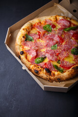 Pizza with cheese, ham, green basil leaves and tomatoes in paper box on black background. Food product advertising banner, copy space