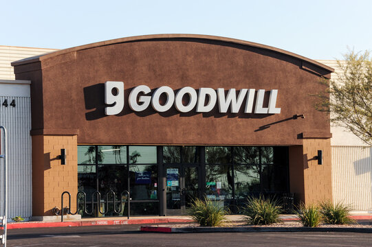 Front entrance of a Goodwill thrift store with a brown facade and clear glass doors, surrounded by minimal desert landscaping under bright morning sun