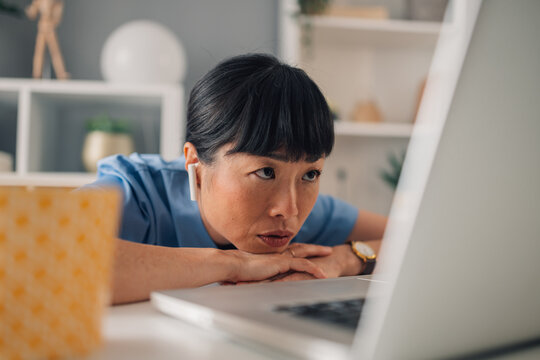 Asian woman nurse in blue scrubs leaning on desk, looking at laptop screen.