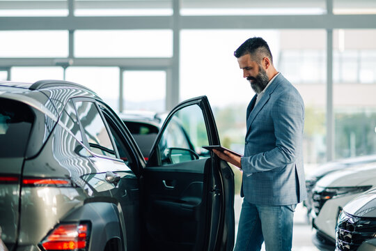 Businessman in suit examining car in dealership with digital device.